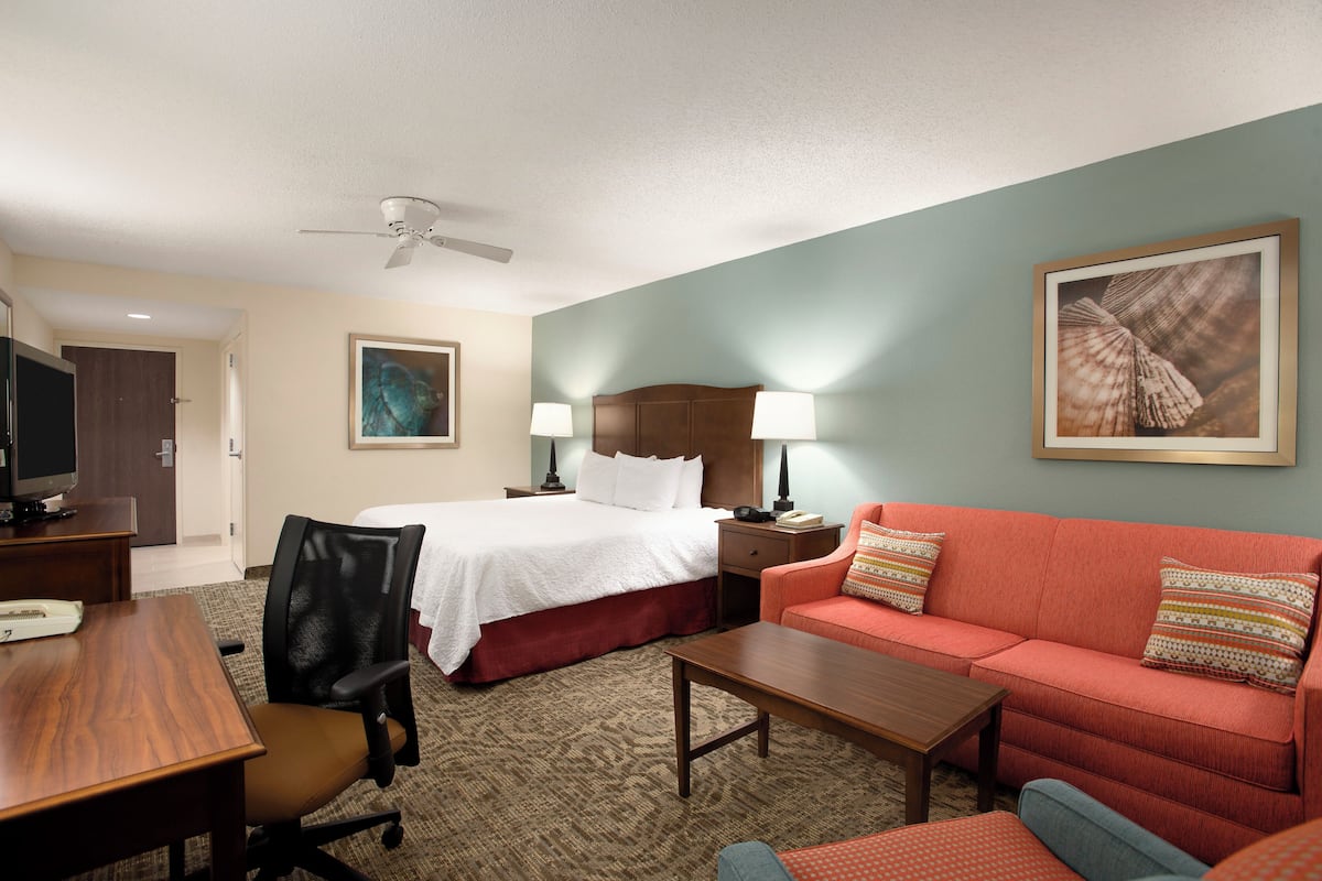 A hotel room with a bed, a red sofa, a wooden table, a desk, a chair, a ceiling fan, and two lamps.