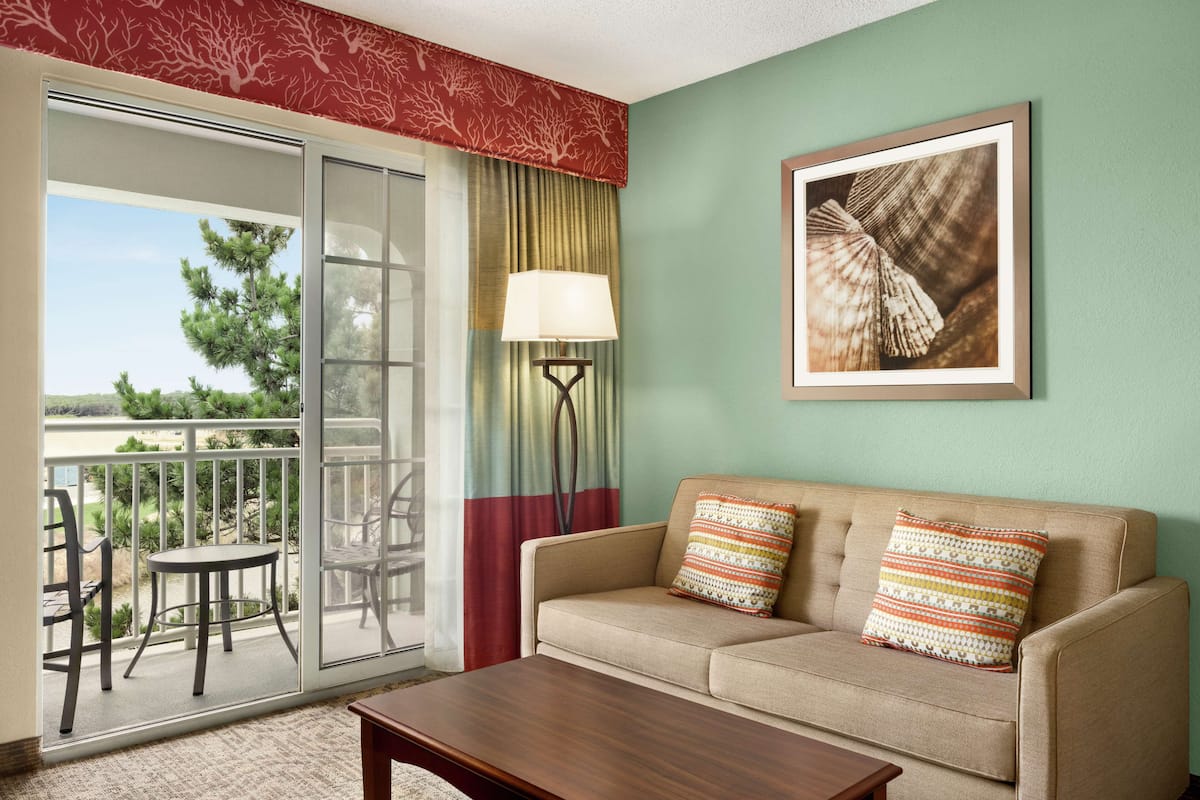 A room with a sofa, a table, a lamp, a framed picture, and a view of a balcony with chairs and a tree.