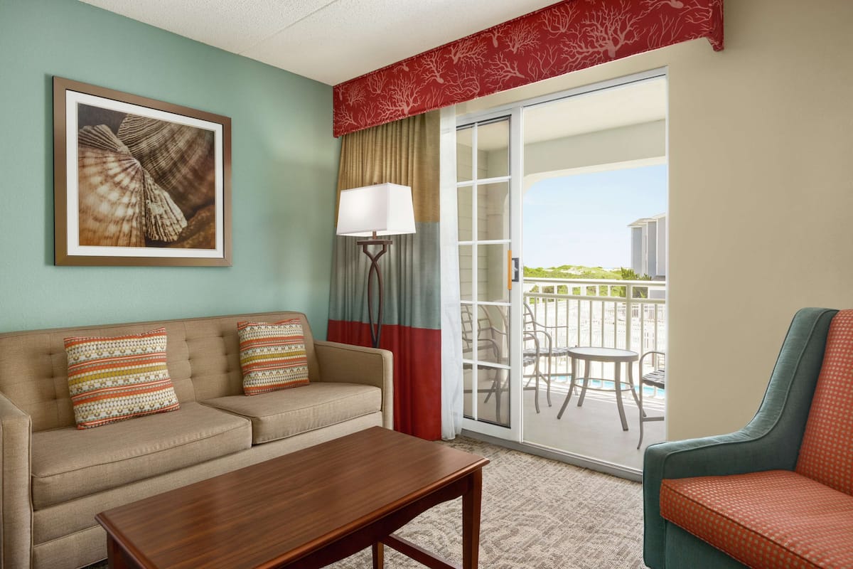 A hotel room with a beige sofa, a wooden coffee table, a patterned armchair, a framed seashore picture, a floor lamp, and a sliding glass door leading to an outdoor area with a pool and chairs.