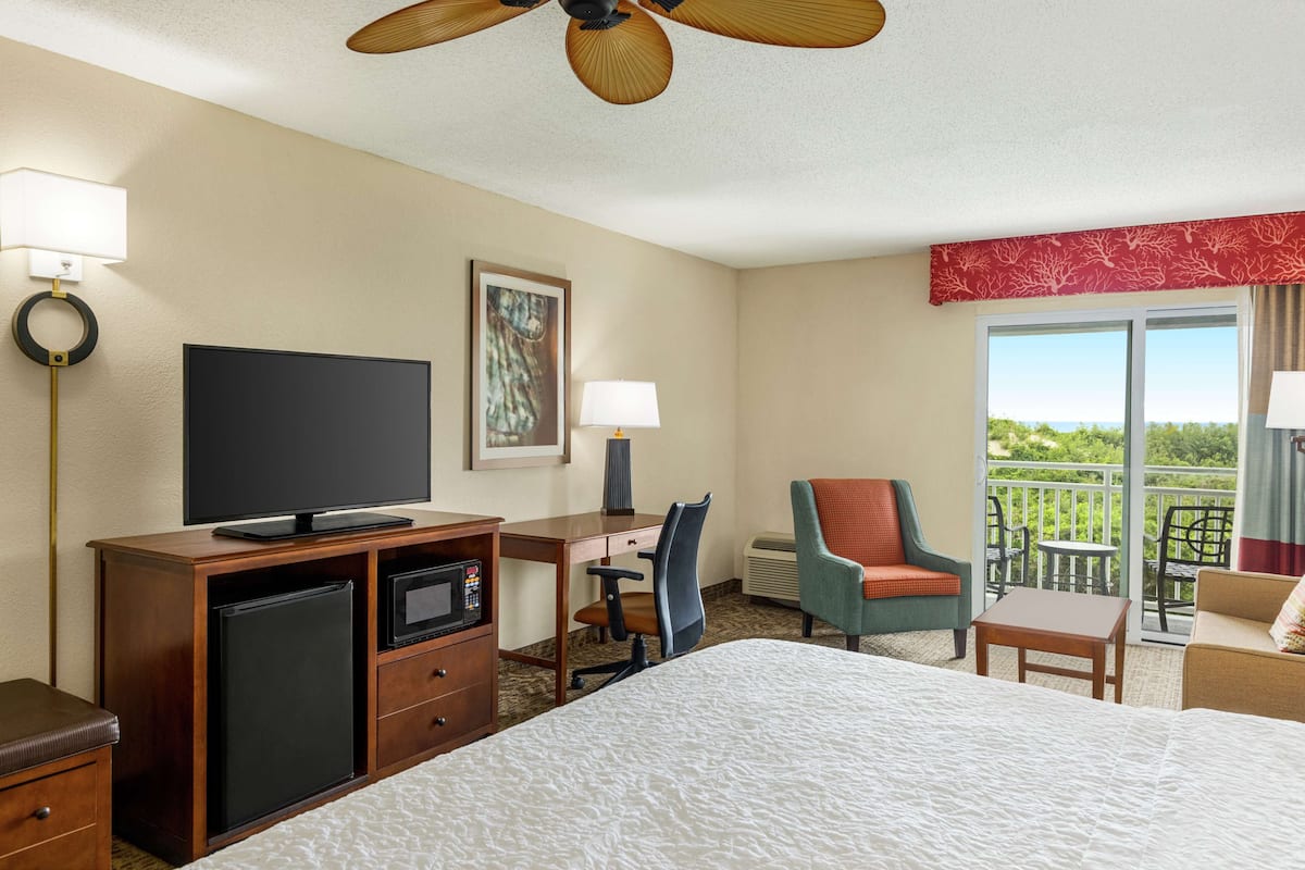 A hotel room with a bed, a desk with a computer, a TV, a small fridge, a chair, and a balcony with a view.