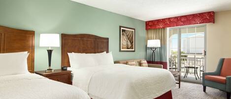 A hotel room with two beds, a sitting area with a chair, a balcony with a view, and a ceiling fan.