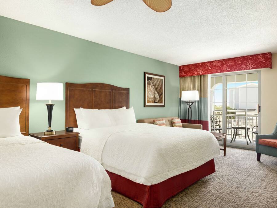 A hotel room with two beds, a sitting area with a chair, a balcony with a view, and a ceiling fan.