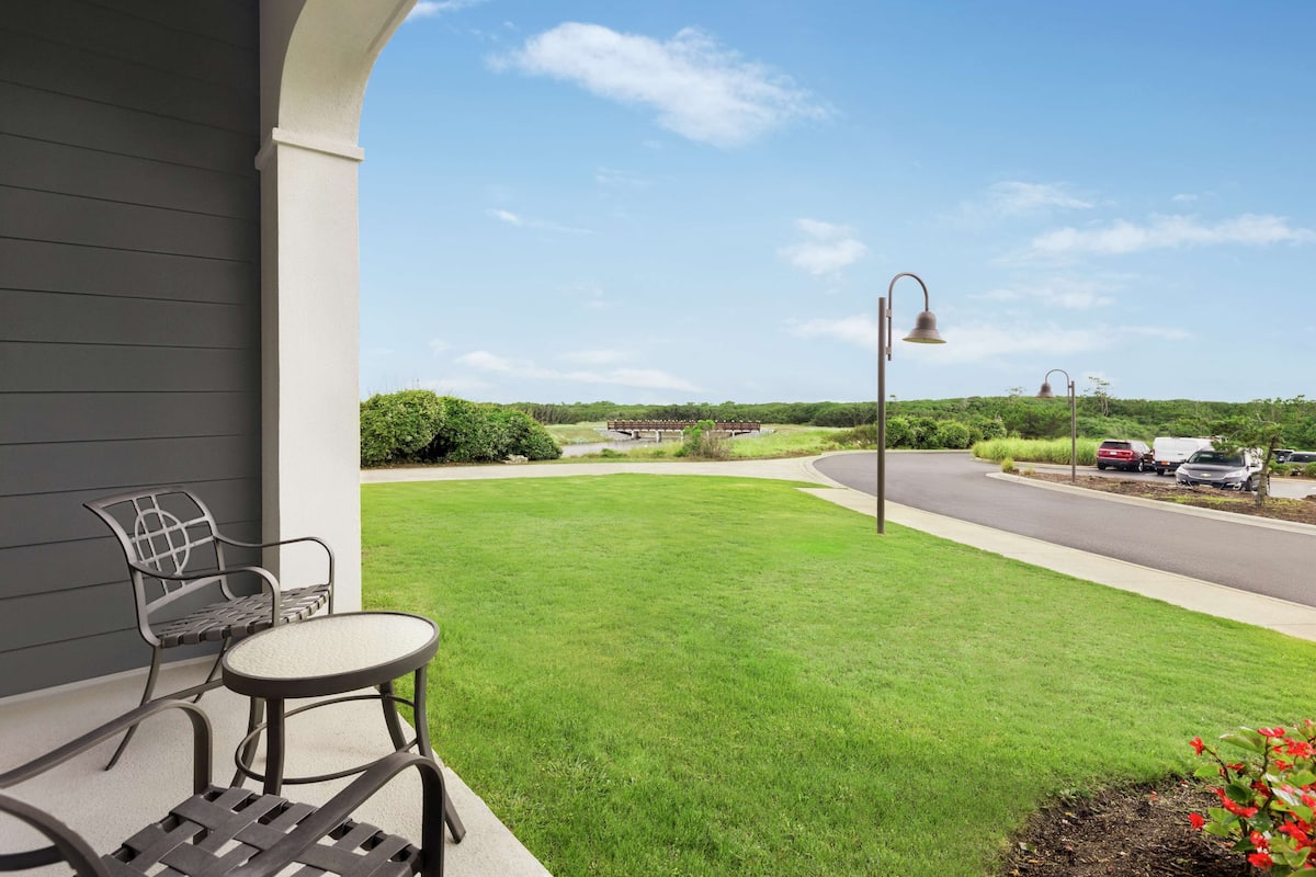 A patio with a table and chairs, a view of a road, and a green lawn.