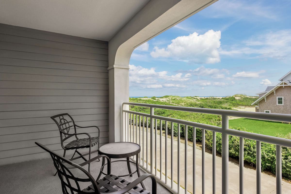 A balcony with a table and chairs, offering a view of a green landscape and a building in the distance.