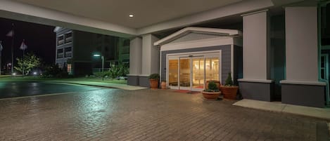 En hotelindgang om natten med en belagt indkørsel, plantekasser og en bygning med en glasdør.