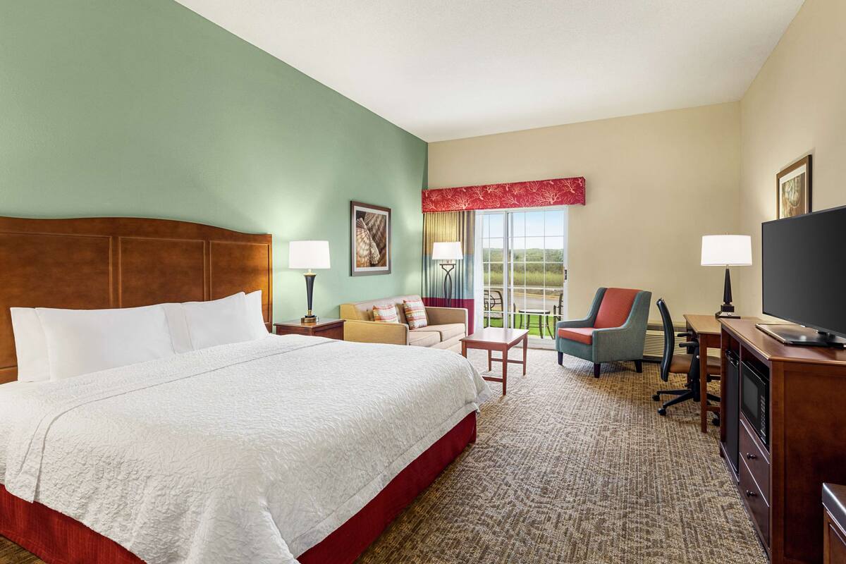 A hotel room with a large bed, a desk with a television, a chair, and a view of the outdoors.