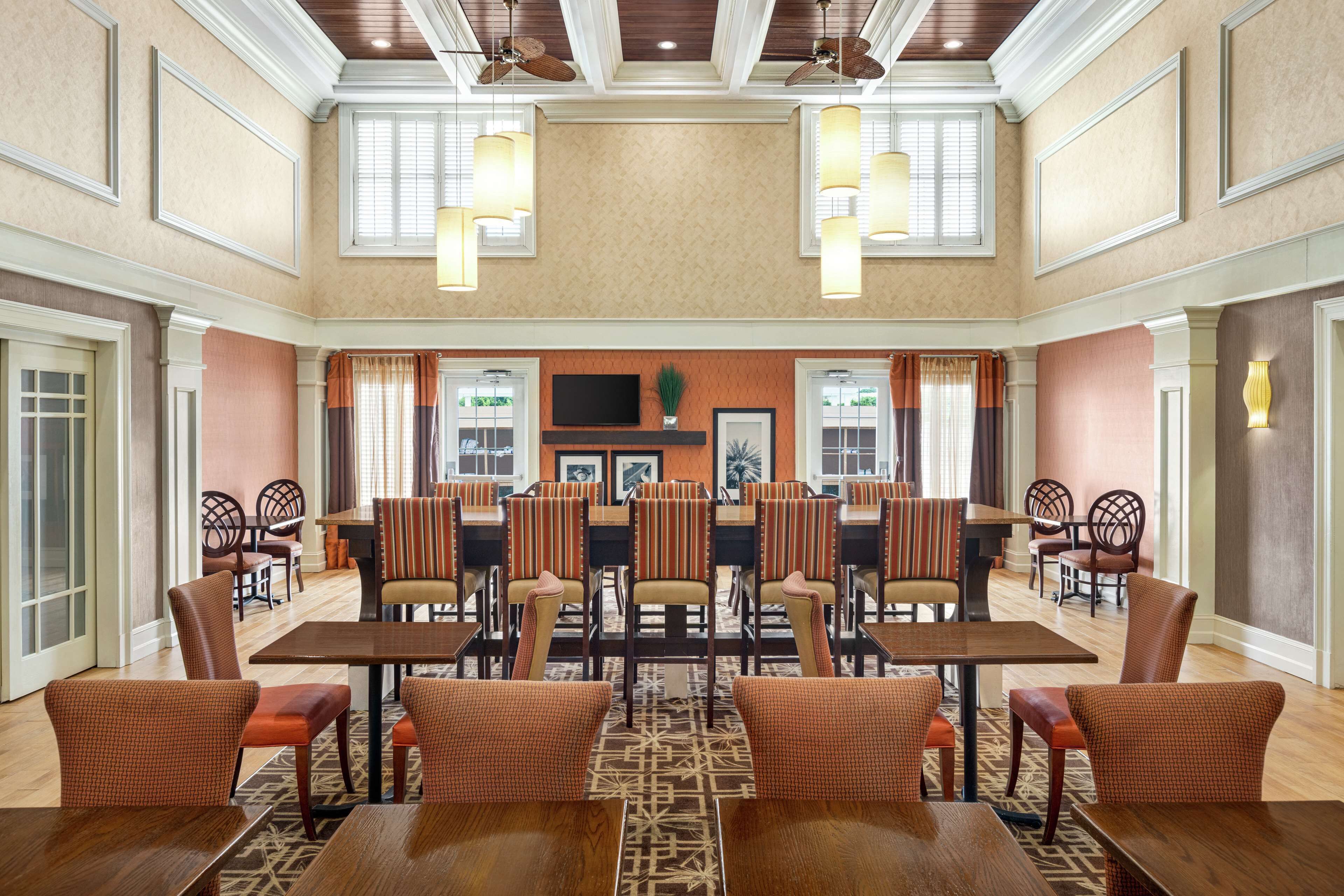 A spacious dining area with multiple tables and chairs, a flat-screen TV mounted on the wall, and ceiling fans.