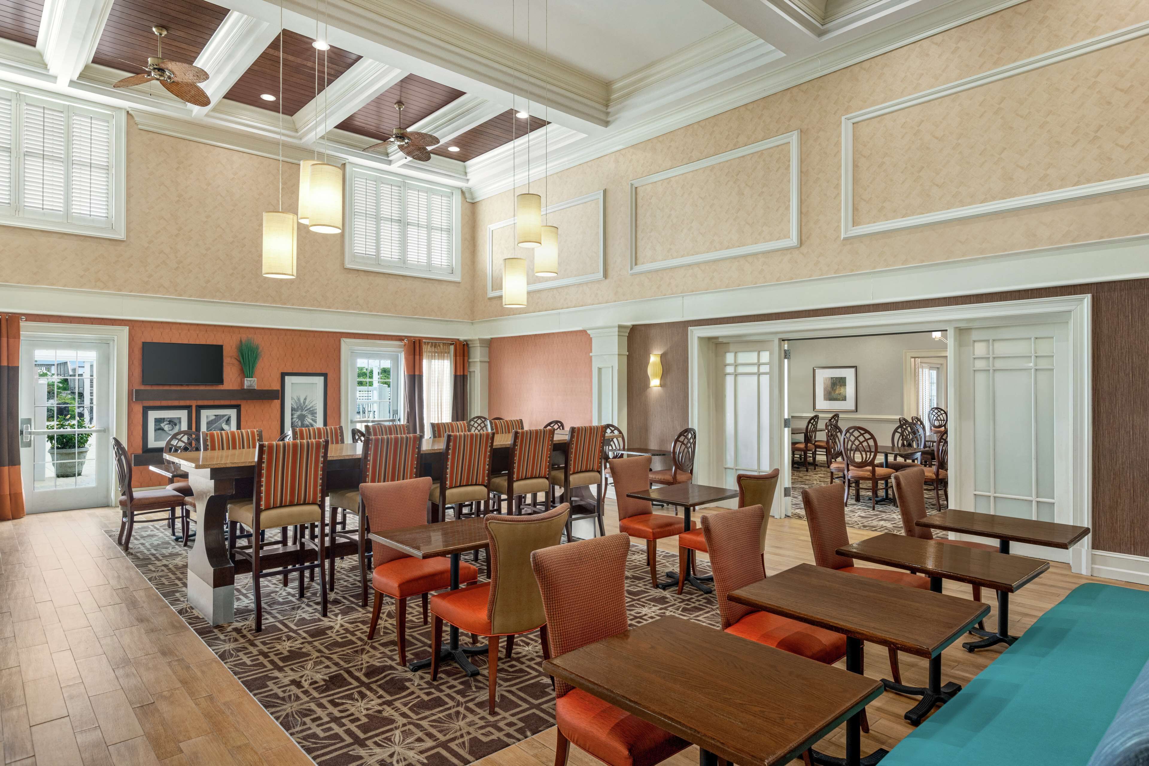 A spacious dining area with multiple tables and chairs, high ceilings with wooden beams, and large windows allowing natural light.