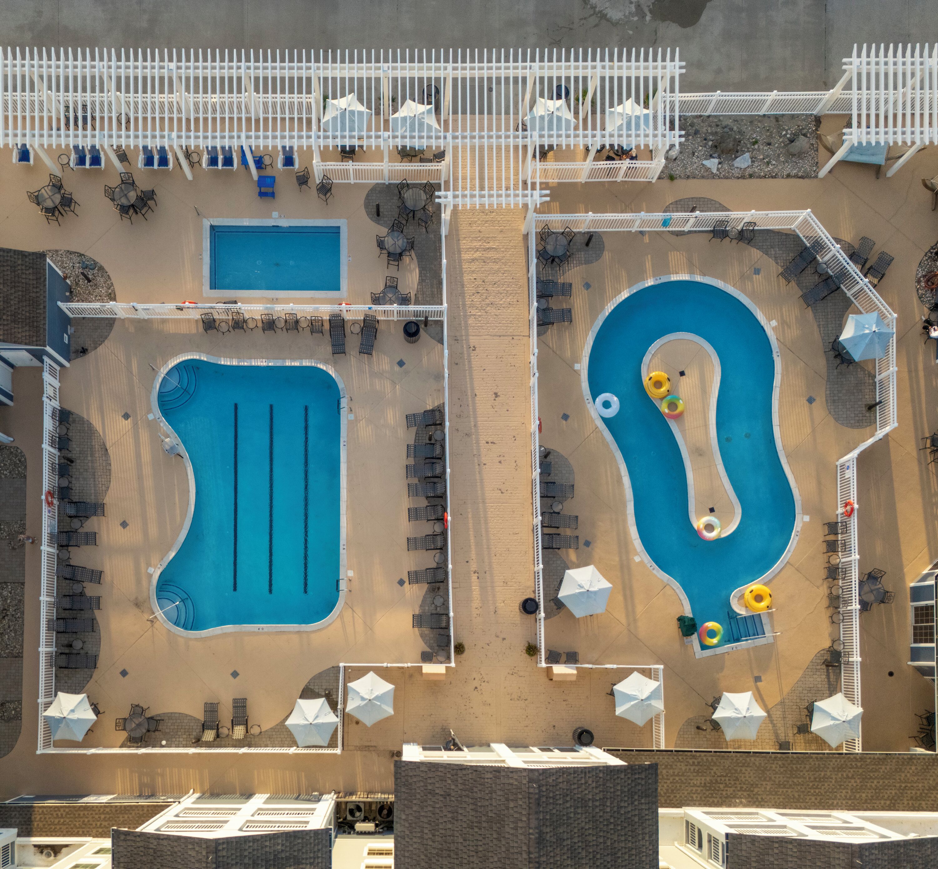 An aerial view of a pool area with two separate pools, sun loungers, umbrellas, and a white fence.