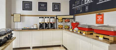 A breakfast bar with coffee makers, a fruit display, and a caution sign for hot surfaces.