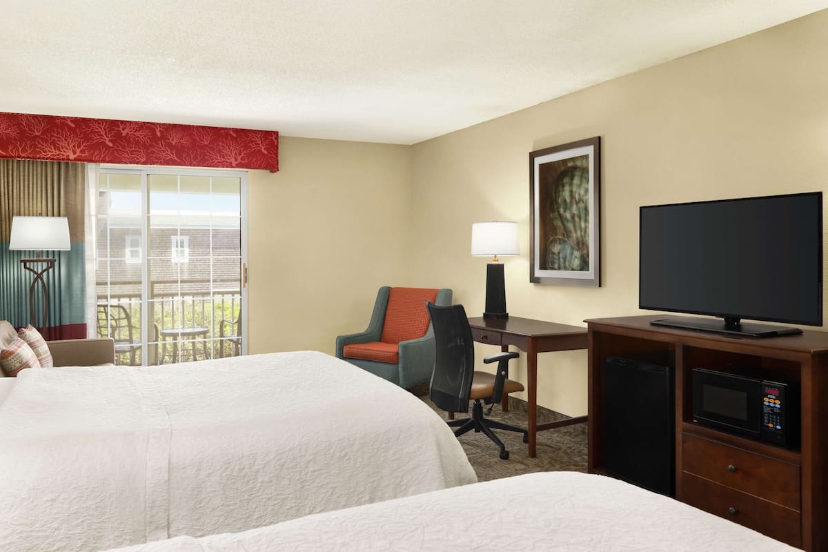 A hotel room with two beds, a desk with a chair, a television, a microwave, and a window with a view of the outside.