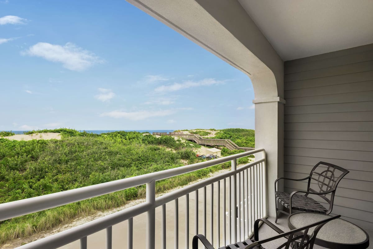 A balcony with a view of a beach and dunes, featuring a metal railing and two chairs.