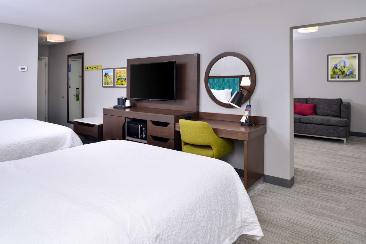 A hotel room with two beds, a desk with a flat-screen TV, a microwave, and a small seating area with a couch and chair.