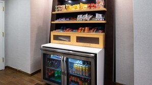 Ein Getränkeautomat mit Snacks und Getränken, ein Kühlschrank mit Getränken und ein Holzregal darüber.