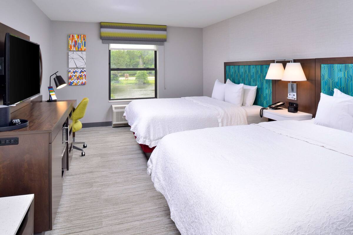 A hotel room with two beds, a desk with a TV, a desk lamp, and a window with a view of greenery.