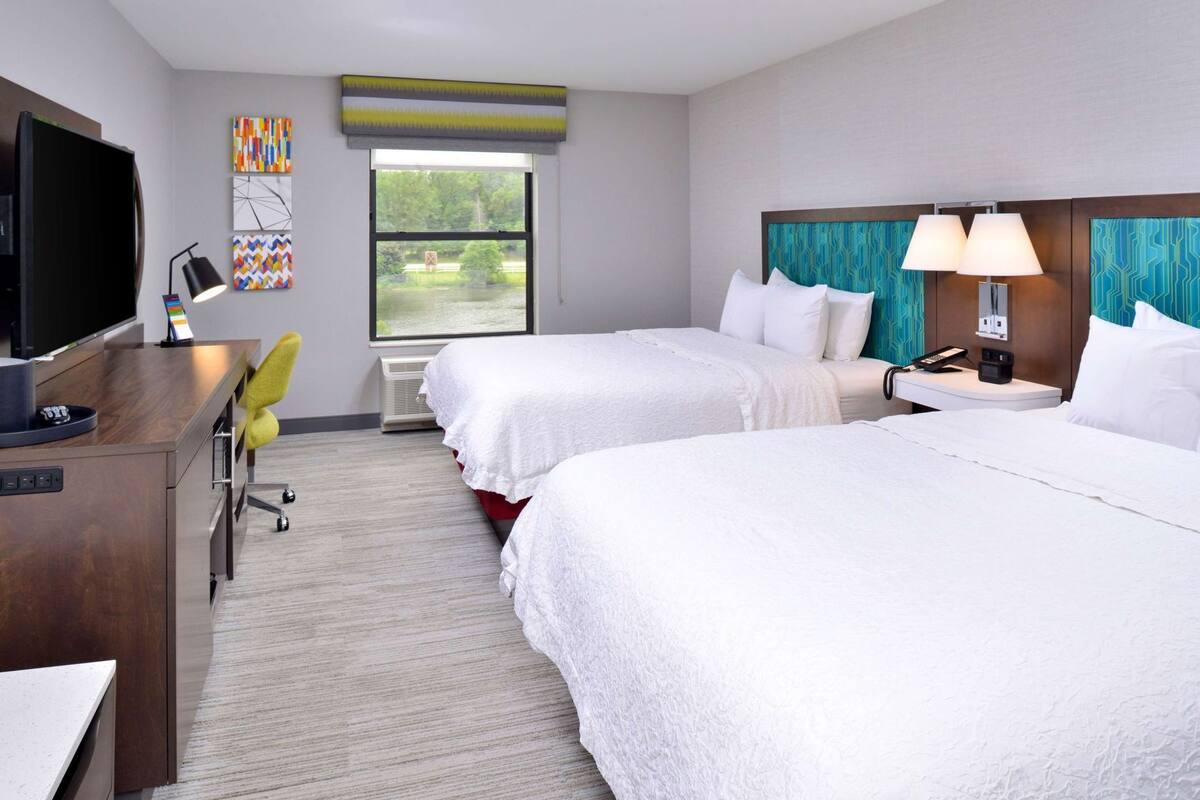 A hotel room with two beds, a desk with a TV, a chair, and a window with a view of trees.