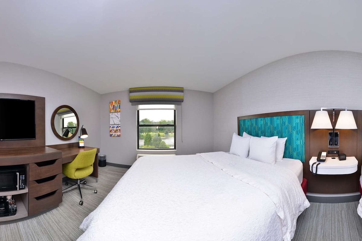 A hotel room with two beds, a desk with a chair, a TV, a mirror, a window, and a bathroom.