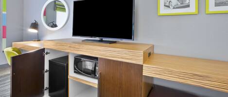 A modern hotel room with a flat-screen TV, a built-in microwave, a wooden bench, and a small seating area.