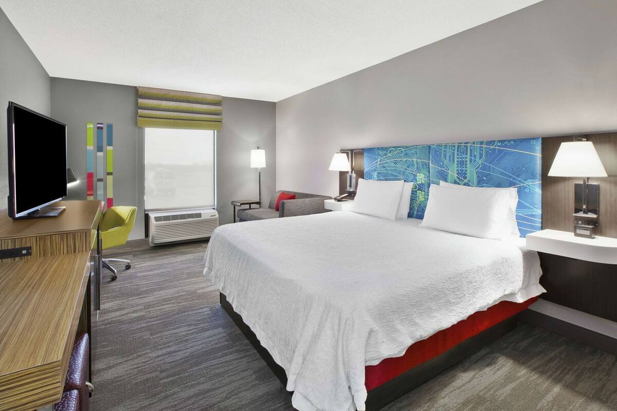 Une chambre d’hôtel avec un grand lit, un bureau doté d’un téléviseur à écran plat, une chaise, une lampe et un mur orné d’un motif représentant un circuit imprimé bleu.