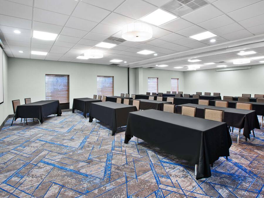 Une salle de conférence équipée de tables et de chaises, d’un écran de projection et de fenêtres avec des stores.