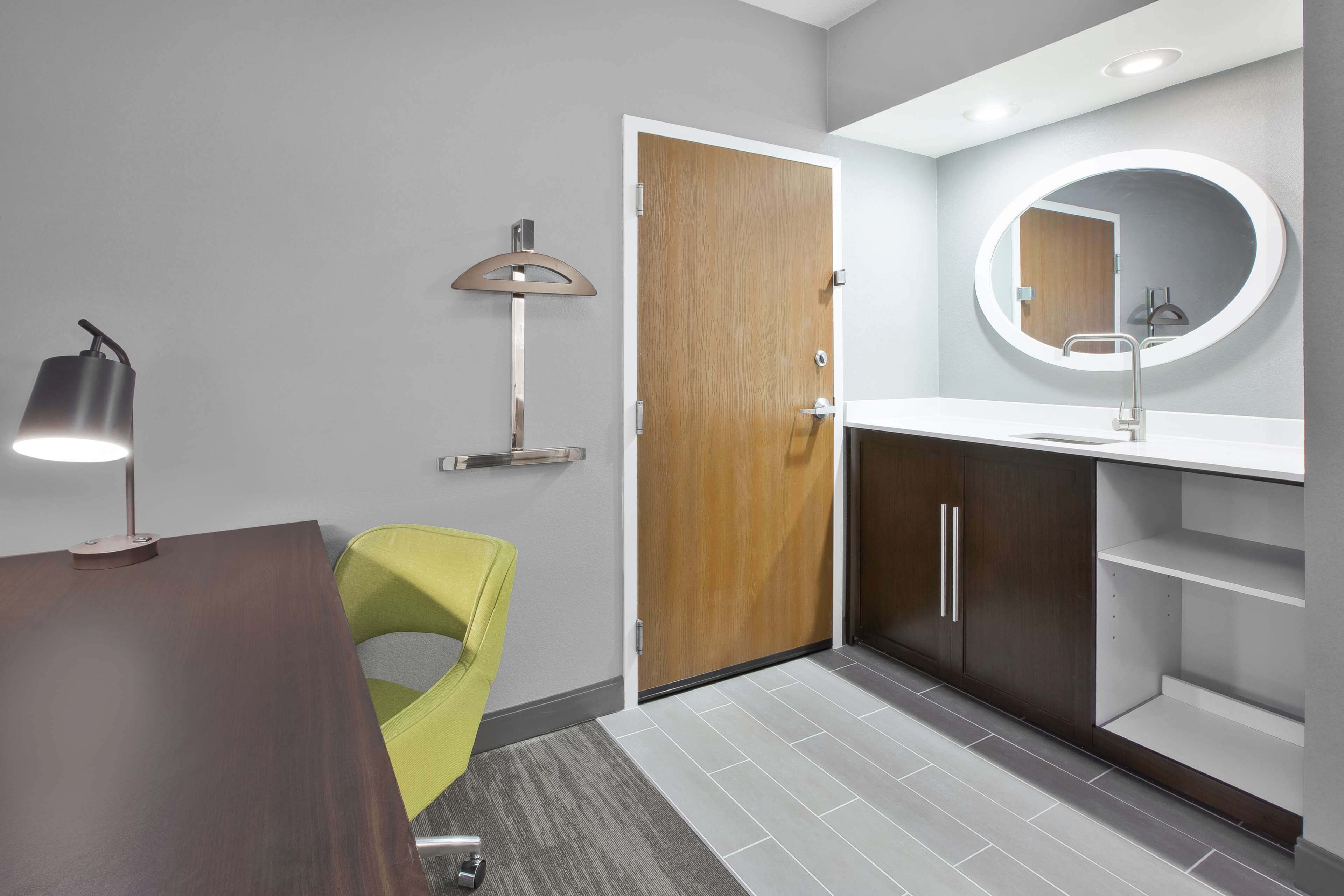A modern bathroom with a wooden door, a round mirror, a white sink, and a green chair.