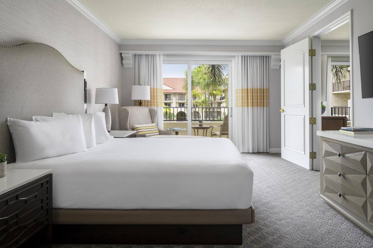 A hotel room with a large bed, a desk with a lamp, a chair, a dresser, and a balcony with a view of palm trees.