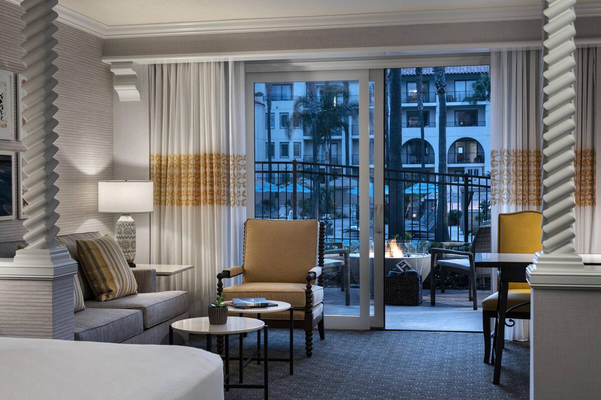 A hotel room with a sofa, armchair, small round table, and a view of the outdoor balcony area.