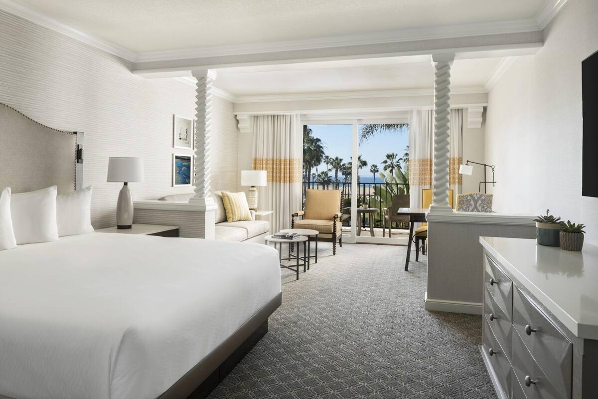 A hotel room with a large bed, a desk, a chair, and a view of palm trees.