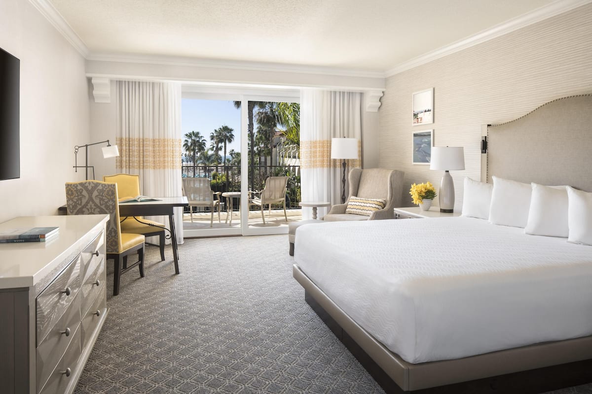 A hotel room with a large bed, a desk with a chair, a small table with a lamp, and a view of the outdoors.