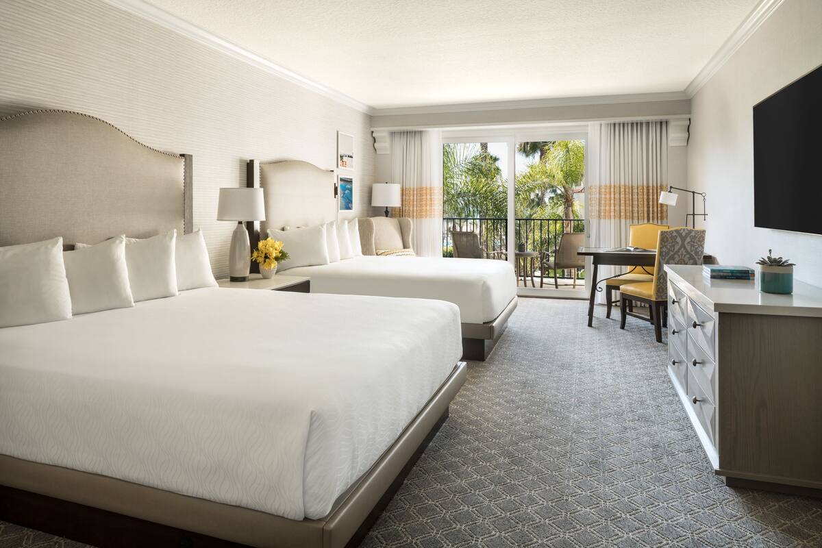 A hotel room with two beds, a desk, a chair, and a balcony with a view of trees.