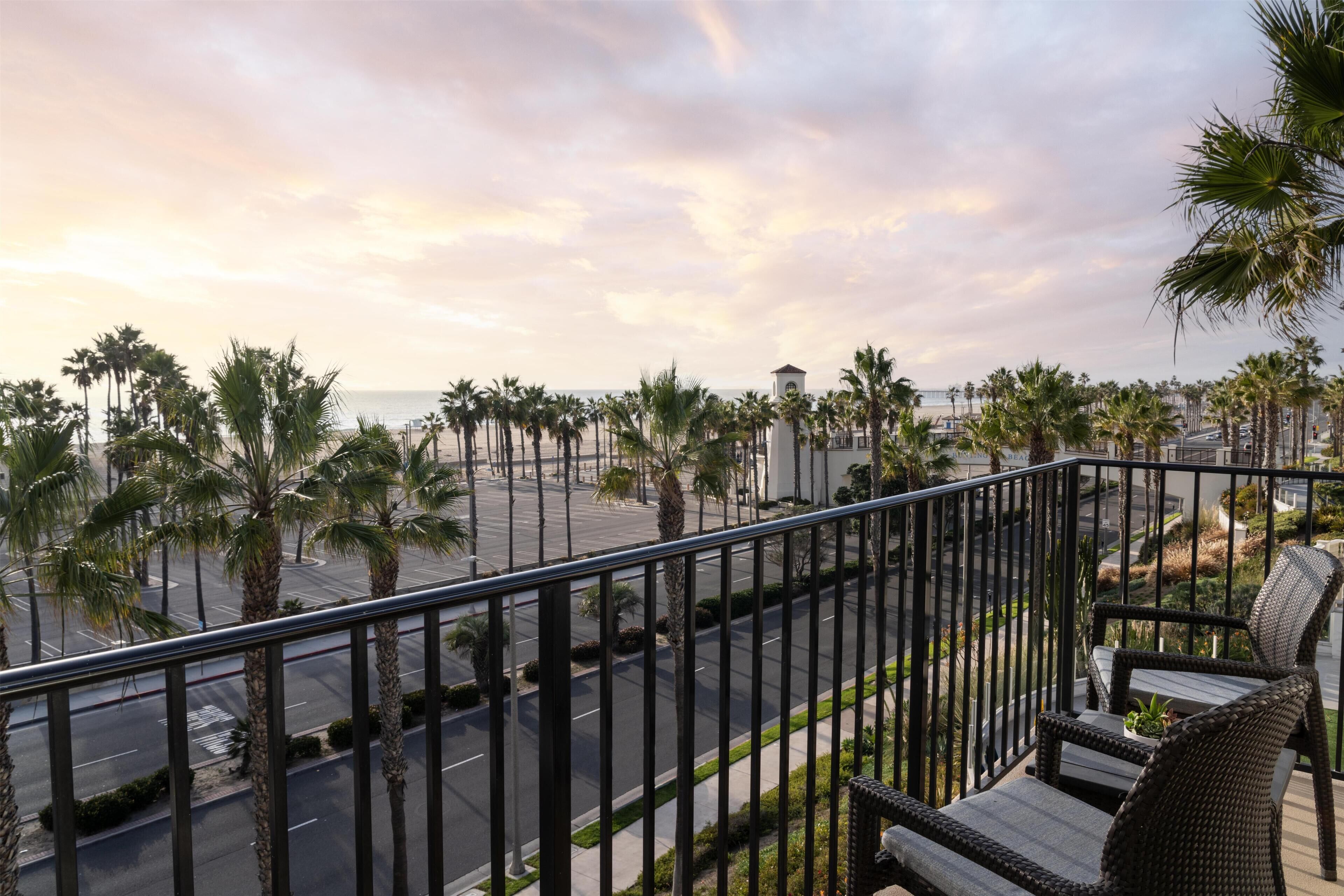 A balcony with wicker furniture overlooking a street lined with palm trees and a distant view of the ocean.