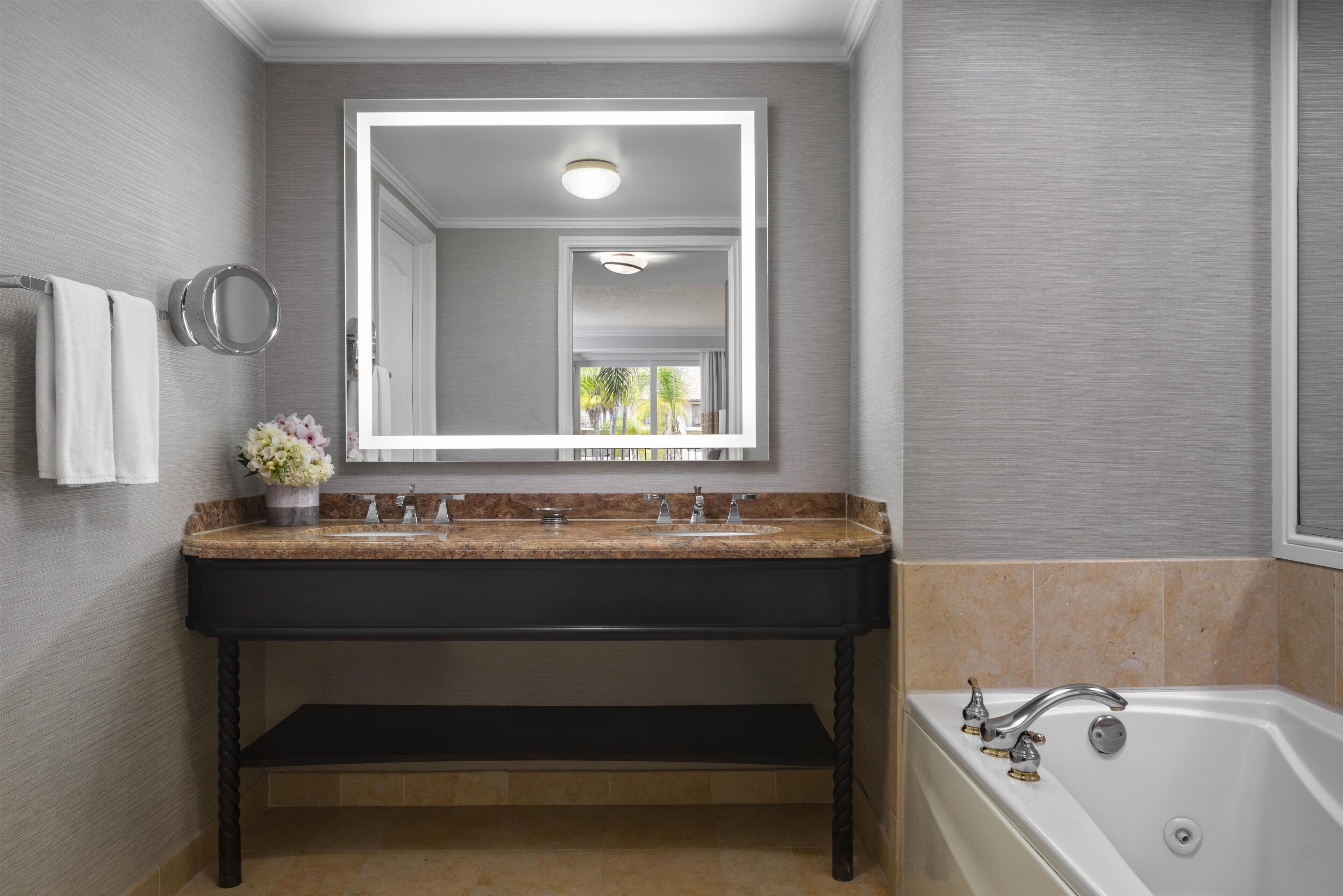 Un baño con lavamanos de encimera de granito, espejo, toallero, tocador y bañera.