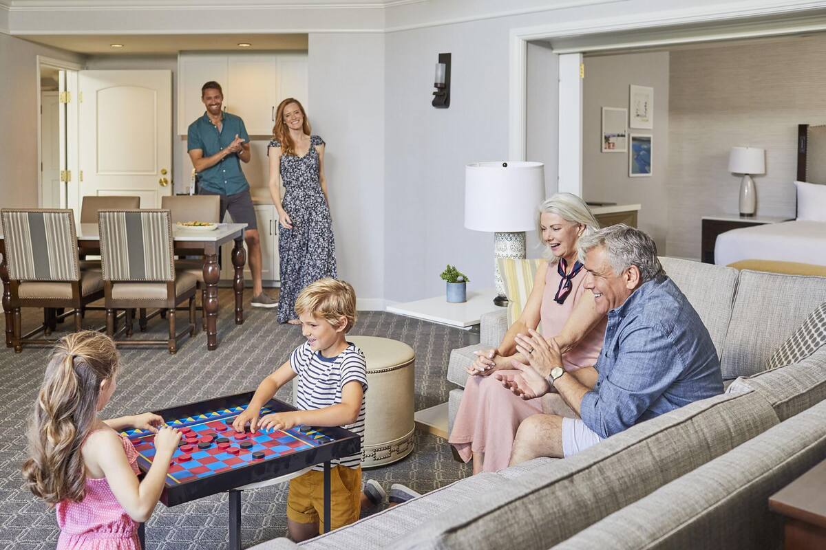 A family playing a board game in a hotel room with a sofa, dining table, and a lamp.