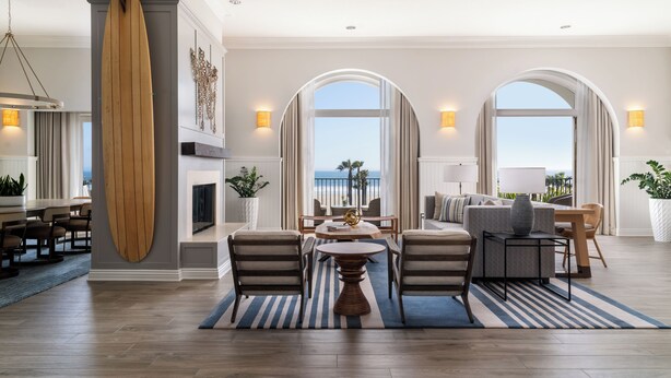 A spacious living room with a large wooden surfboard, a fireplace, a dining area, and a view of the ocean through arched windows. - Hyatt Regency Huntington Beach Resort and Spa (Huntington Beach)