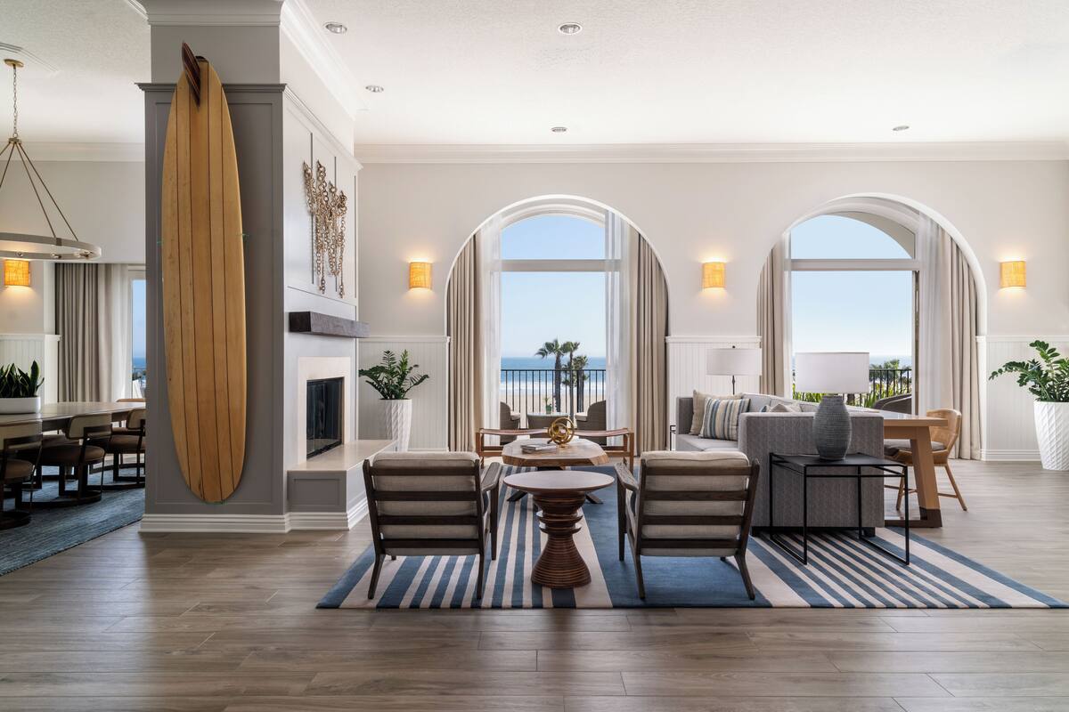 A spacious living room with a large wooden surfboard, a fireplace, a dining area, and a view of the ocean through arched windows.