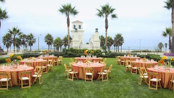 A formal outdoor event setup with round tables and chairs, palm trees, and a white building in the background.