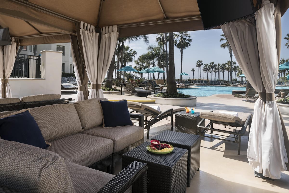 A covered outdoor seating area with wicker furniture, a pool, and palm trees in the background.