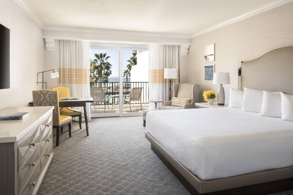 A hotel room with a large bed, a desk with a chair, a TV, and a balcony with a view of palm trees and the ocean.
