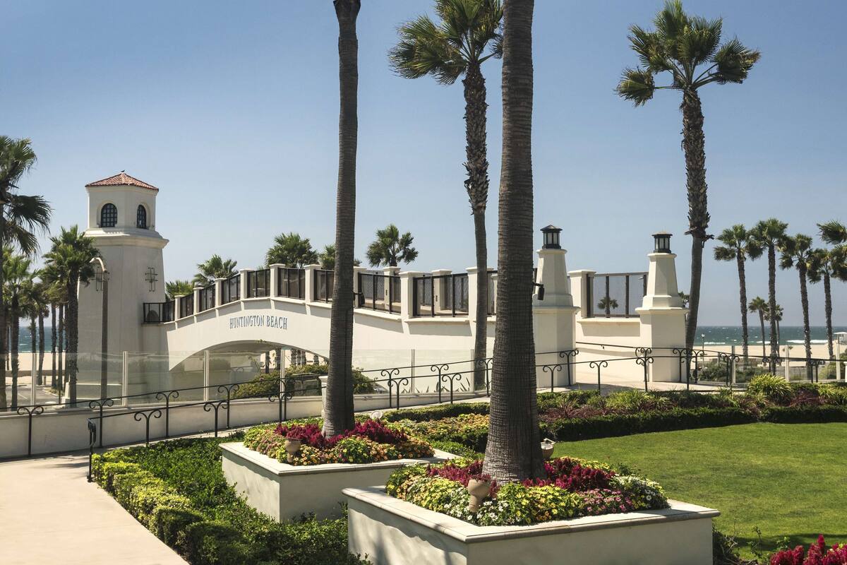 A white building with a tower and a bridge, surrounded by palm trees and well-maintained gardens.