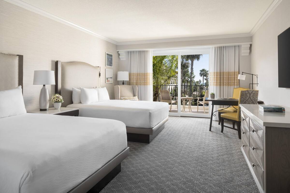 A hotel room with two beds, a desk, a chair, a TV, and a balcony with a view of palm trees.