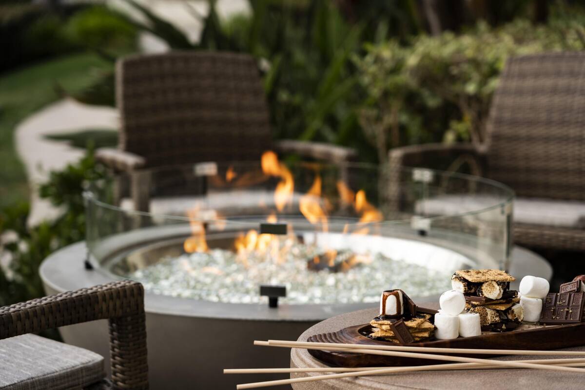 A fire pit surrounded by wicker chairs, with marshmallows and chocolate bars on a tray.