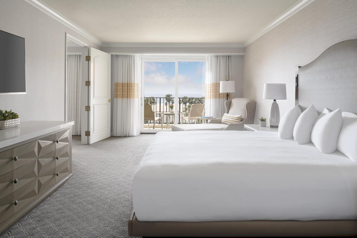 A hotel room with a large bed, a desk, a chair, a balcony with a view, and a television.