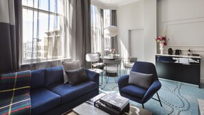A modern hotel room with a blue sofa, a matching armchair, a coffee table, a dining area with a small table and chairs, and a view of the city through large windows. - Fable Dunedin (Dunedin)