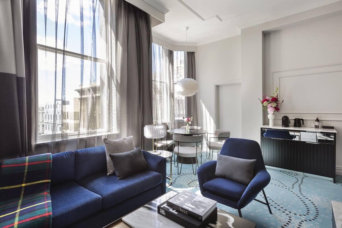A modern hotel room with a blue sofa, a matching armchair, a coffee table, a dining area with a small table and chairs, and a view of the city through large windows.