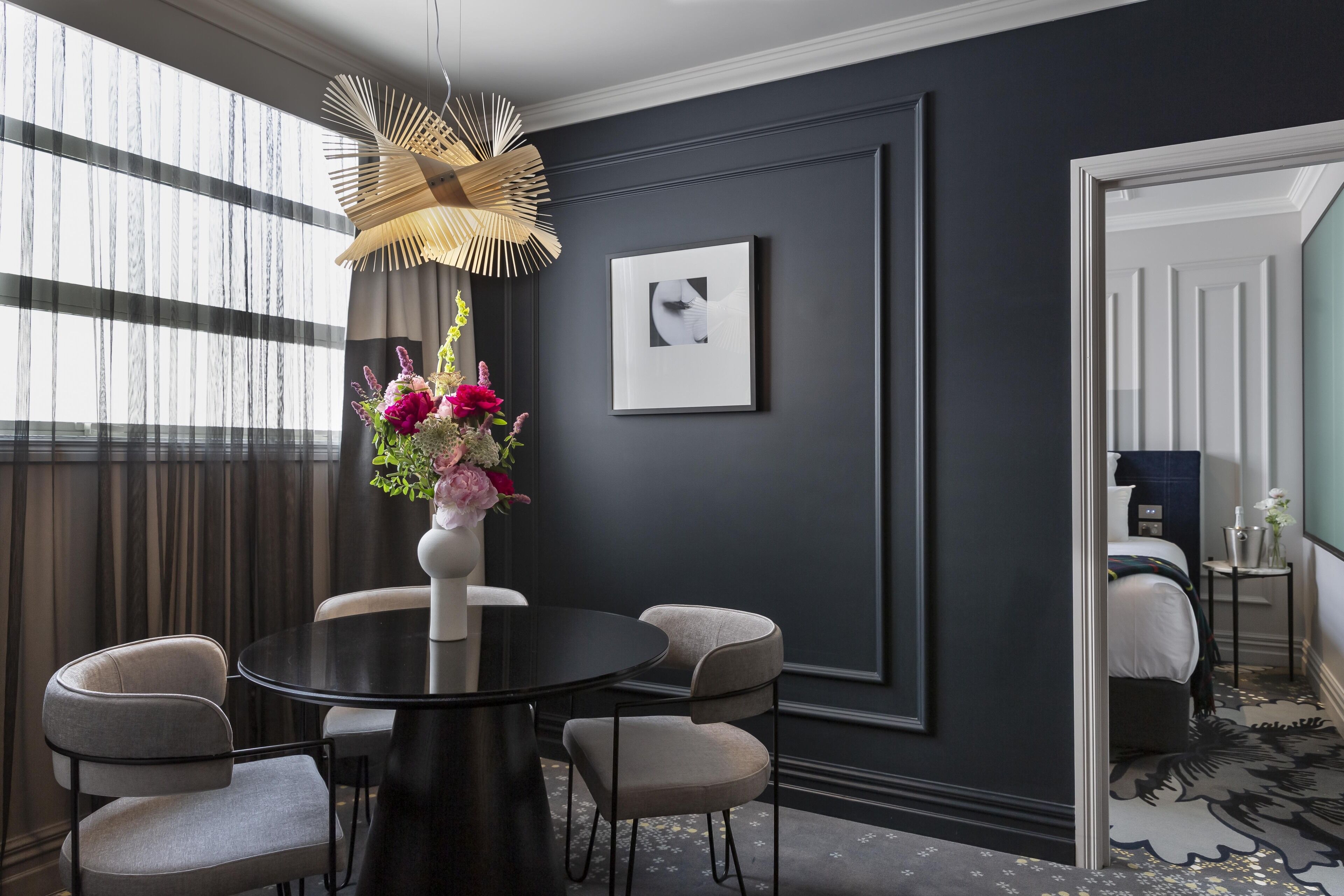A modern room with a round table, four chairs, a vase with flowers, a framed picture on the wall, and a bed in the background.
