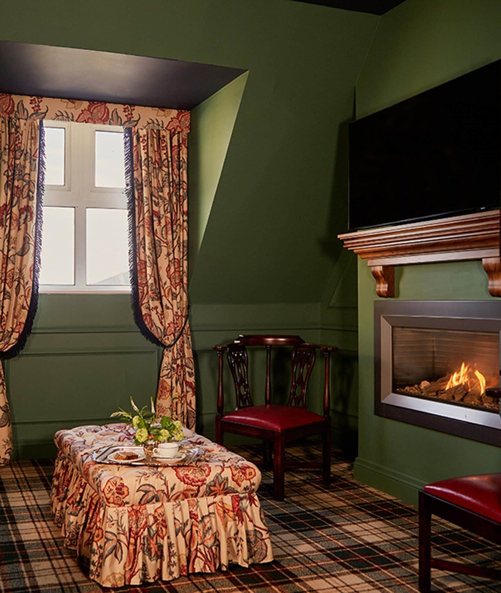 A cozy room with a plaid floor, a table with a floral tablecloth, a chair, a fireplace, and a large window with curtains.