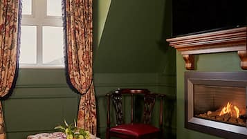 A cozy room with a plaid floor, a table with a floral tablecloth, a chair, a fireplace, and a large window with curtains.
