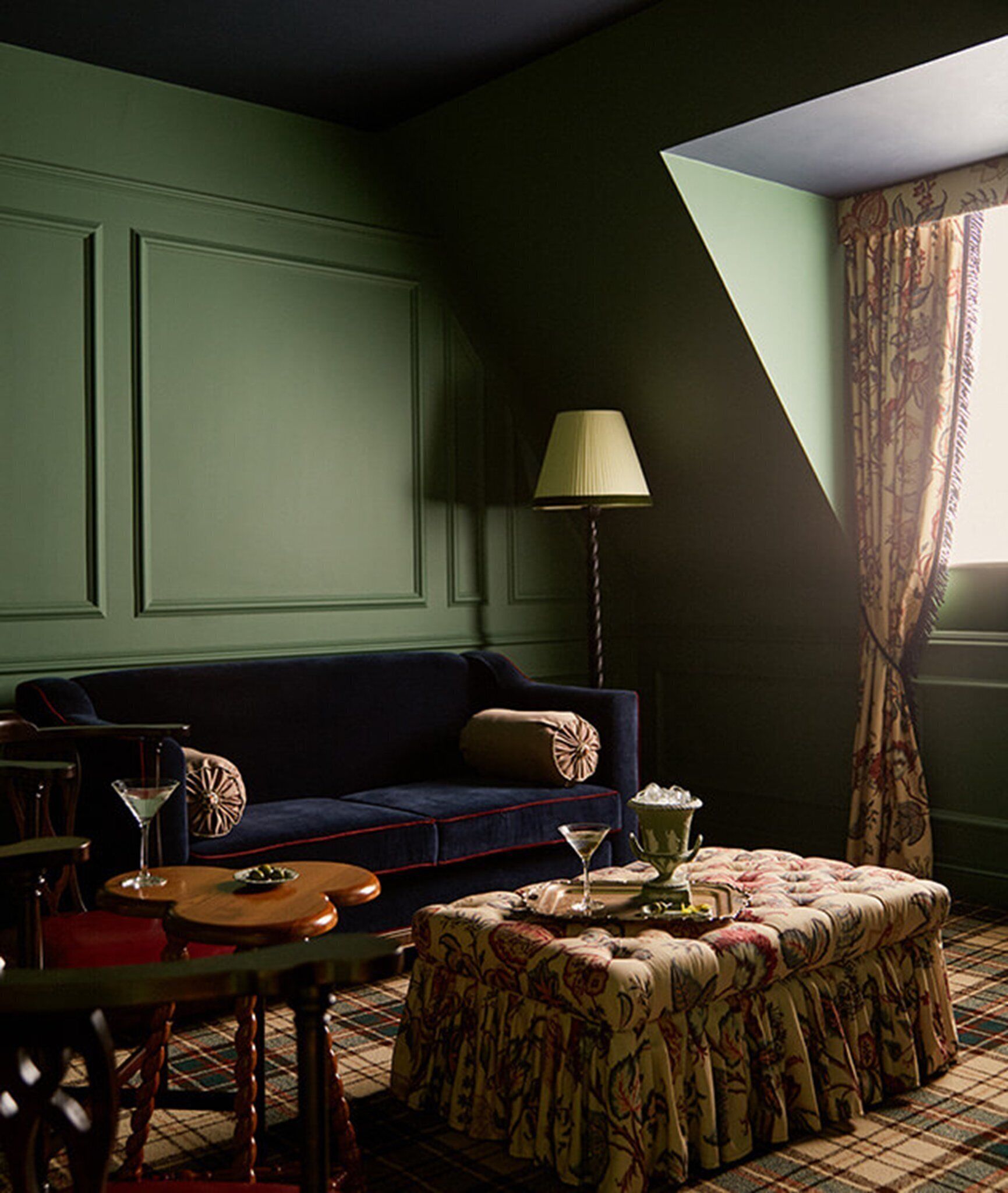 Una habitación con pared verde oscuro, un sofá azul, un otomano estampado, una lámpara de pie y una alfombra de cuadros.