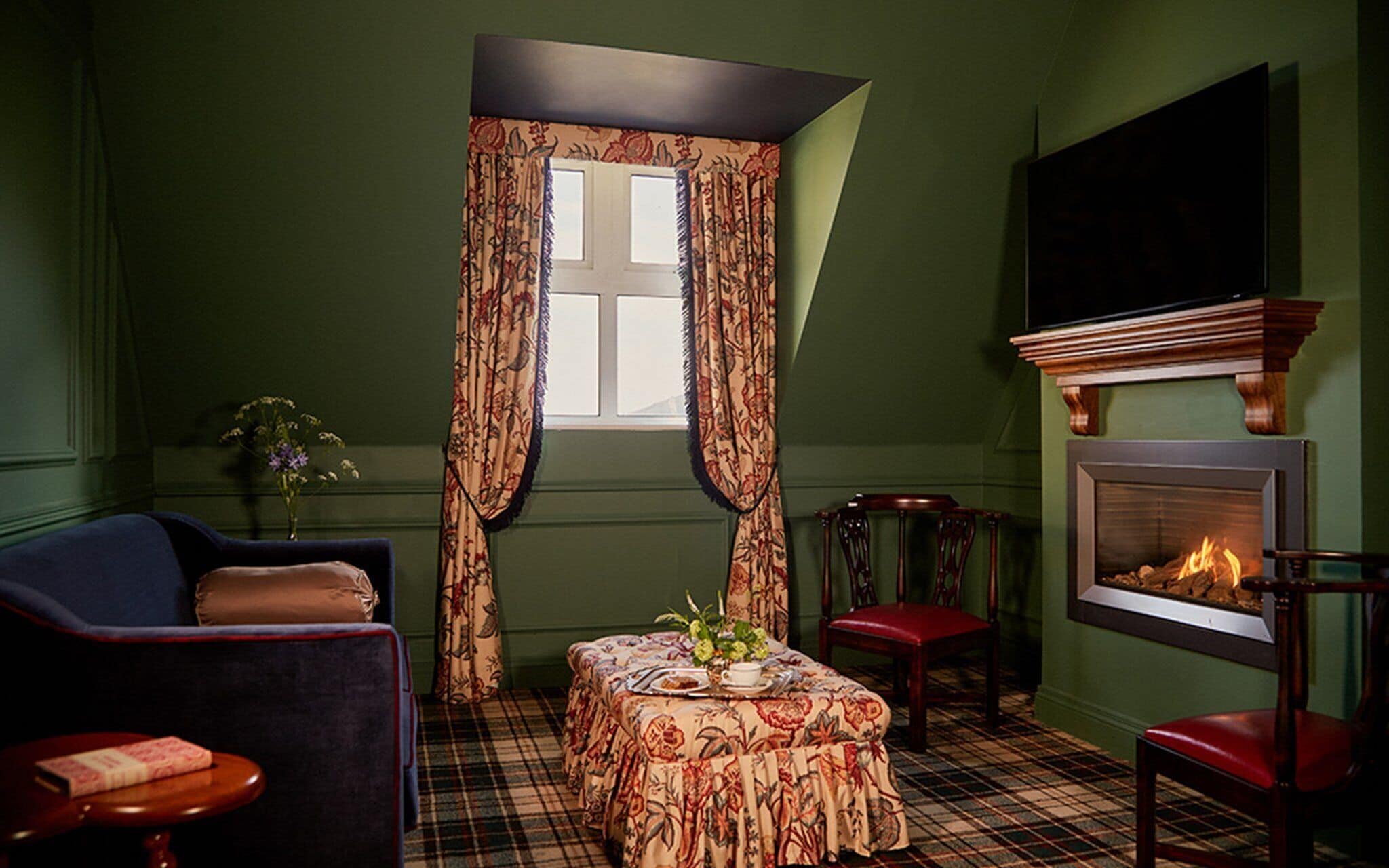 Een gezellige kamer met een geruit vloerkleed, een open haard, een televisie en een raam met bloemen gordijnen.