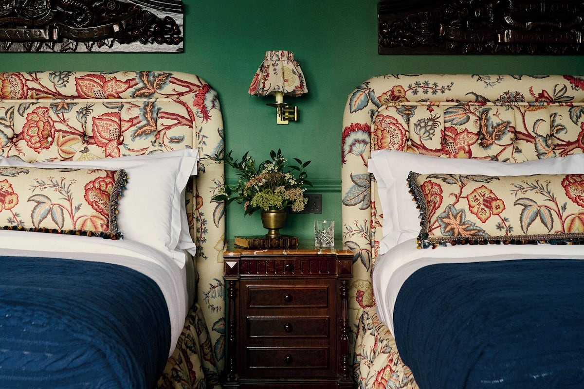 A room with two beds, each with floral-patterned bedding and headboards. There is a small wooden nightstand with a lamp and a vase of flowers between the beds.